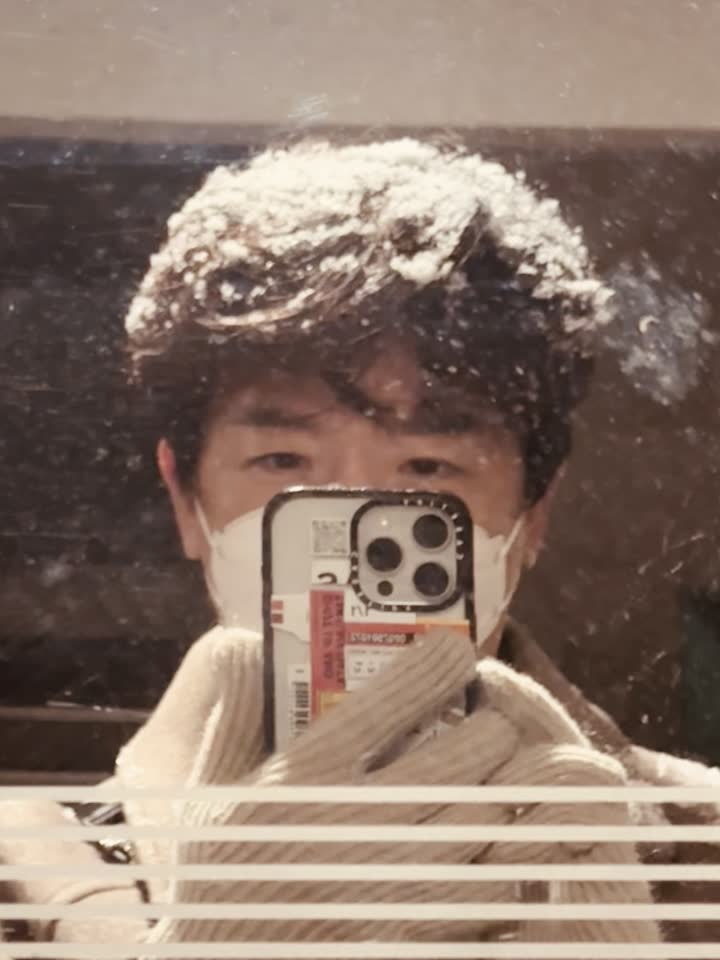 My reflection in a glass door, phone held up in front of my face, snow sitting unmelted across the top of my hair.